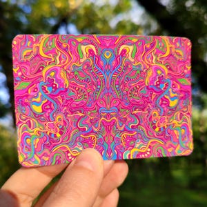 May include: A rectangular card with a vibrant, psychedelic design. The artwork features a complex pattern of swirling lines and shapes in shades of pink, yellow, blue, and green. The card is held in a person's hand, with a blurred green background.