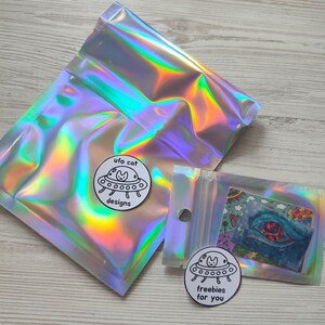 May include: Two iridescent cellophane bags with a holographic effect. One bag is sealed, and the other is open, revealing a small card. Two circular stickers with a cat in a UFO design are also visible. The text on the stickers reads "ufo cat designs" and "freebies for you."