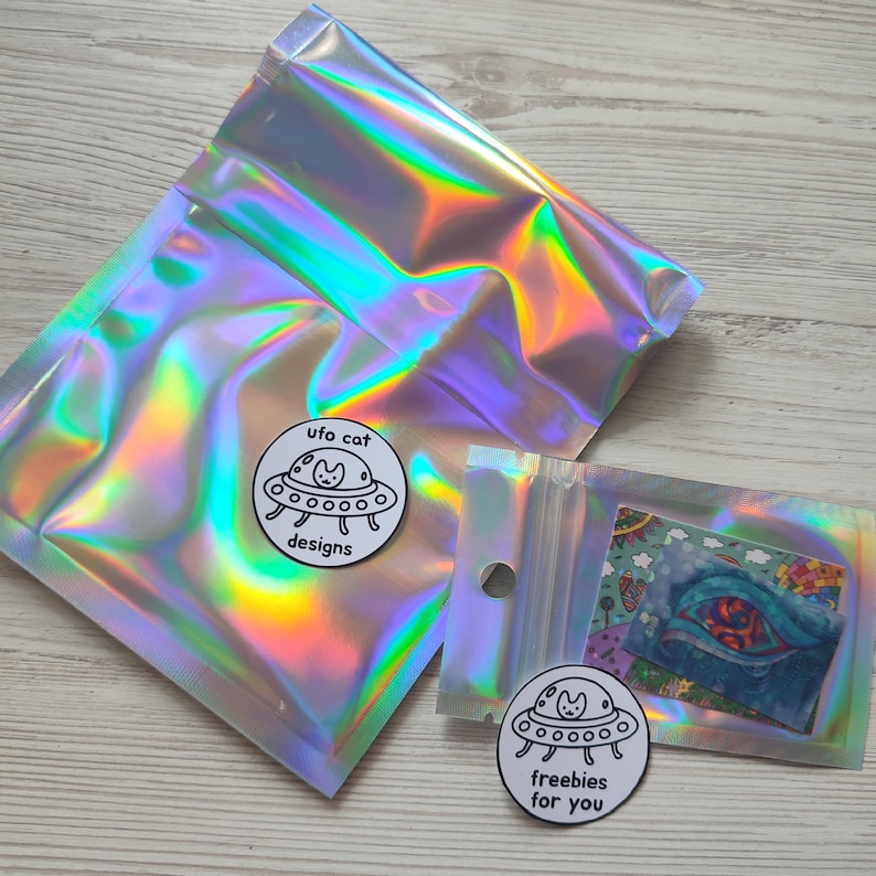 May include: Two iridescent cellophane bags with a holographic effect. One bag is sealed, and the other is open, revealing a small card. Two circular stickers with a cat in a UFO design are also visible. The text on the stickers reads "ufo cat designs" and "freebies for you."