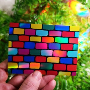 May include: A rectangular card featuring a vibrant brick wall design. The bricks are a mix of bright colors including red, yellow, blue, pink, and green, outlined in black. The card is held against a backdrop of green foliage.