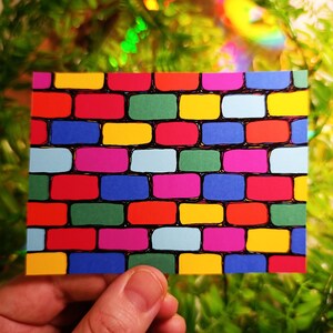 May include: A colorful brick pattern design on a rectangular card. The bricks are in various colors, including red, green, yellow, blue, and pink, with black outlines. The card is held in a person's hand against a blurred green background.