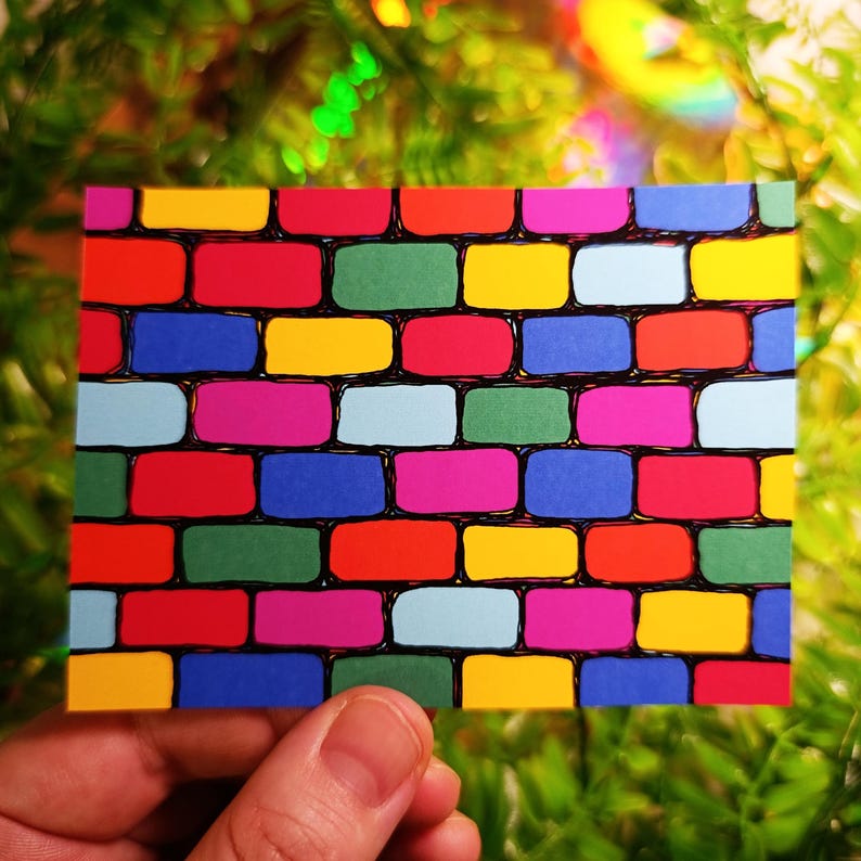 May include: A colorful brick pattern design on a rectangular card. The bricks are in various colors, including red, green, yellow, blue, and pink, with black outlines. The card is held in a person's hand against a blurred green background.