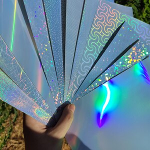 May include: A hand holding a fan of light blue holographic paper sheets. Each sheet has a different pattern, including dots, swirls, and geometric designs. The paper reflects a rainbow of colors, creating a shimmering effect.