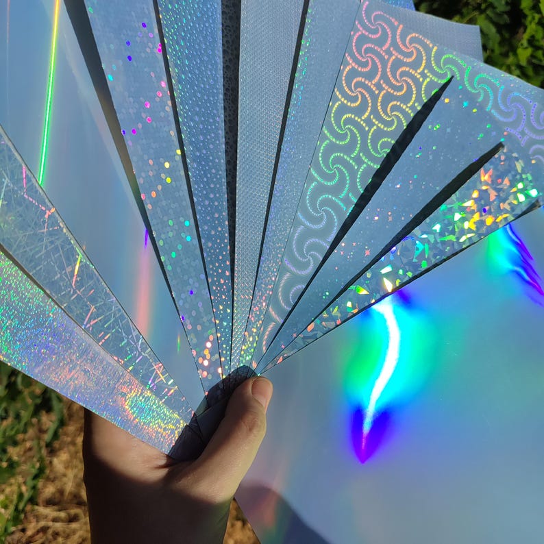 May include: A hand holding a fan of light blue holographic paper sheets. Each sheet has a different pattern, including dots, swirls, and geometric designs. The paper reflects a rainbow of colors, creating a shimmering effect.