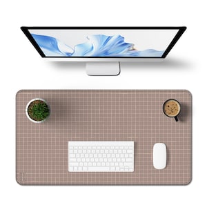Op de afbeelding: Een computeropstelling met een grote monitor met een blauw en wit abstract ontwerp. Een bruine bureaumat met een rasterpatroon bevat een wit toetsenbord, muis, een kleine plant in een pot en een kopje koffie. De opstelling is strak en modern.