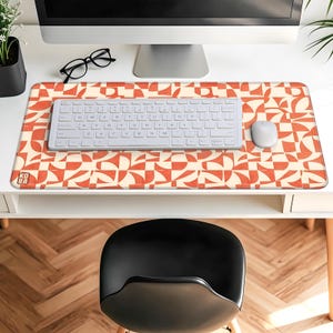 May include: A rectangular desk mat featuring a retro geometric design in cream and orange. A white keyboard and mouse are on the mat, with a computer monitor and glasses in the background. The mat has a logo in the lower left corner.