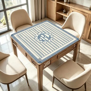 K&ouml;nnte beinhalten: Ein quadratischer Holztisch mit blau-wei&szlig; gestreifter Tischplatte und einem Sonnenemblem. Der Tisch hat vier Beine und eine Schublade. Vier beige St&uuml;hle umgeben den Tisch. Der Tisch steht in einem hellen Raum mit Fenster und Holzschr&auml;nken.