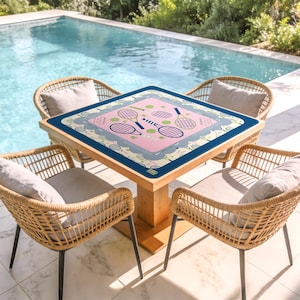 May include: A square wooden table with a pink tennis-themed design, surrounded by four wicker chairs with gray cushions. The table's design features tennis rackets and balls. The scene is set outdoors near a pool.