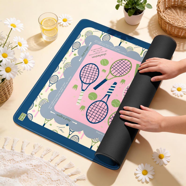 May include: A tennis-themed play mat with a navy blue border and a pink center featuring tennis rackets and balls. The mat is partially rolled up, revealing a black underside. The words "walls &bull; play" are printed on the mat.
