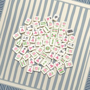 K&ouml;nnte beinhalten: Eine Ansammlung wei&szlig;er Mahjong-Steine, die auf einer gestreiften Oberfl&auml;che verstreut sind. Jede Kachel zeigt chinesische Schriftzeichen, Symbole und rosa Rosenillustrationen. Die Spielsteine sind in einem zuf&auml;lligen Muster angeordnet.