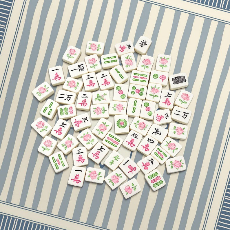 K&ouml;nnte beinhalten: Eine Ansammlung wei&szlig;er Mahjong-Steine, die auf einer gestreiften Oberfl&auml;che verstreut sind. Jede Kachel zeigt chinesische Schriftzeichen, Symbole und rosa Rosenillustrationen. Die Spielsteine sind in einem zuf&auml;lligen Muster angeordnet.