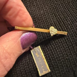 May include: A gold-toned barrette with a heart-shaped cluster of clear rhinestones. The barrette is held near a small tag that reads "MADE IN U.S.A." The background is a dark, textured fabric.