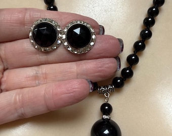 May include: A necklace featuring a strand of round black beads and a large black spherical pendant with a silver-toned bail, accompanied by a pair of round clip-on earrings with faceted black centers surrounded by a border of small clear stones.