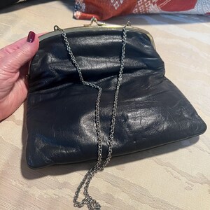 Pu&ograve; includere: Una pochette in pelle nera con una tracolla a catena argentata. La pochette ha una chiusura dorata e una texture leggermente stropicciata.