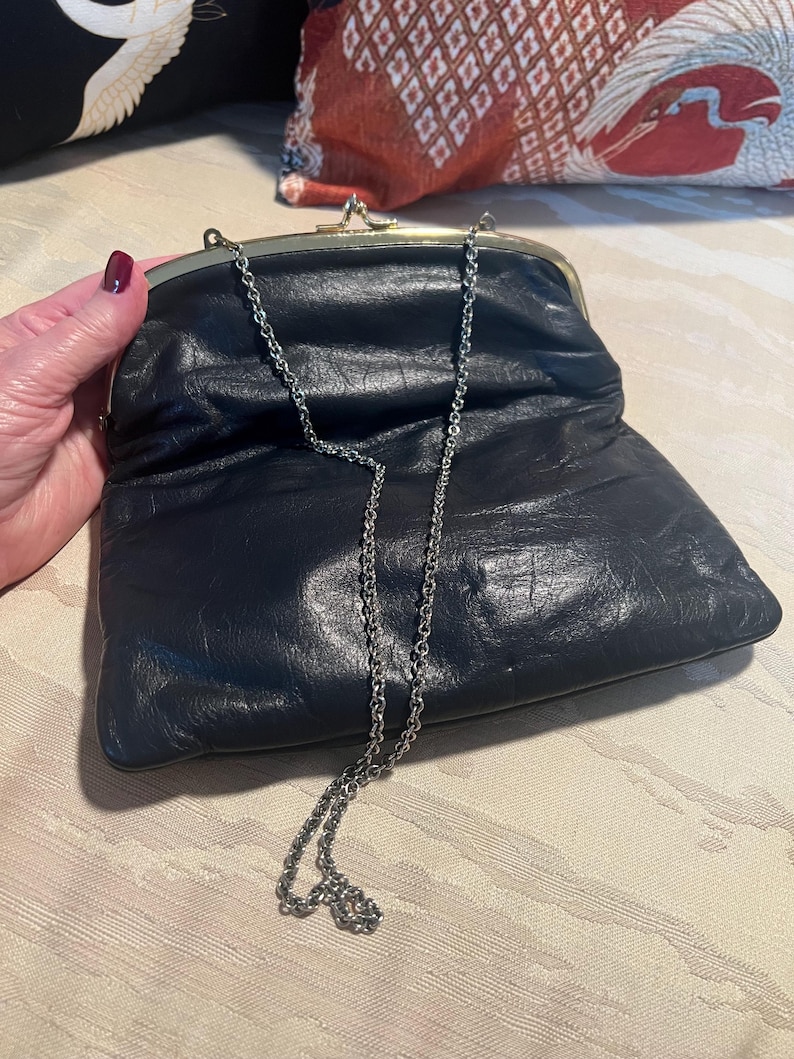 Pu&ograve; includere: Una pochette in pelle nera con una tracolla a catena argentata. La pochette ha una chiusura dorata e una texture leggermente stropicciata.