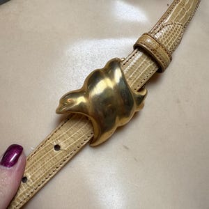 Può includere: Una cintura in pelle beige con fibbia color oro. La cintura presenta un motivo strutturato, simile alla pelle di rettile. La fibbia è un design astratto stilizzato. La cintura è mostrata su un manichino.