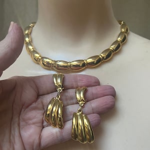 May include: A gold-tone necklace and earrings set. The necklace is a choker style with oval-shaped links. The earrings are dangle style with three curved, overlapping pieces.