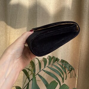 May include: A black velvet clutch with a gold interior. The rectangular bag is held open, revealing the inside. The background is a neutral color, with a plant in the foreground.