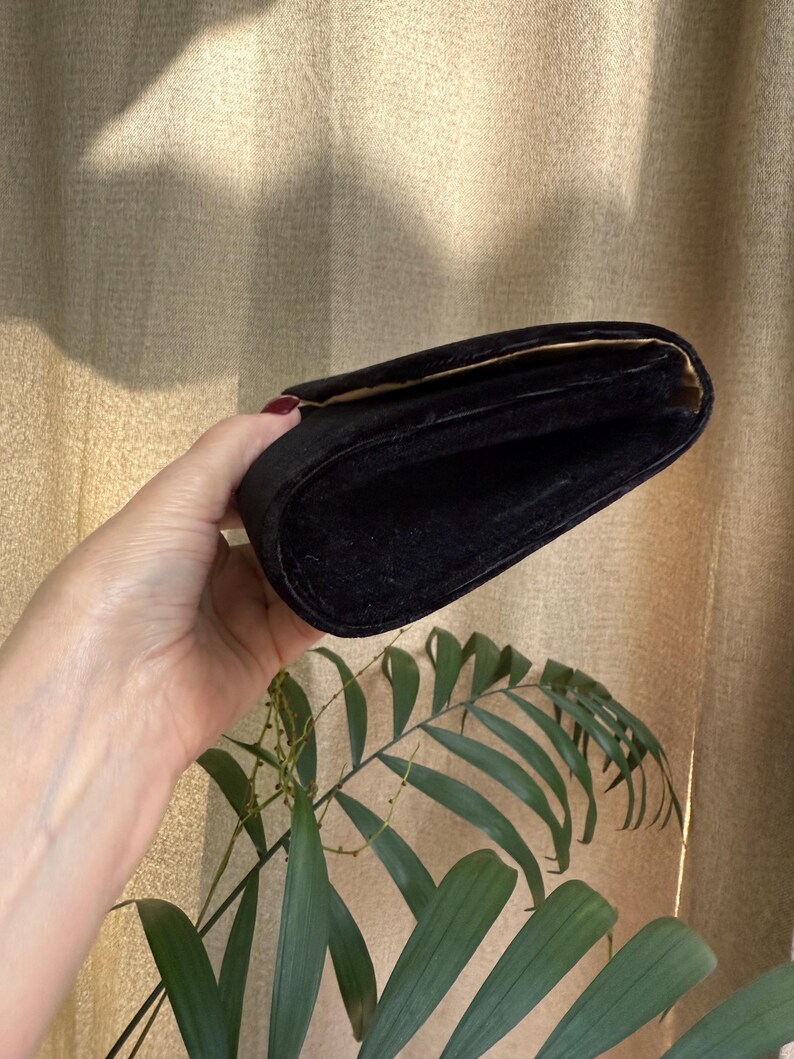 May include: A black velvet clutch with a gold interior. The rectangular bag is held open, revealing the inside. The background is a neutral color, with a plant in the foreground.