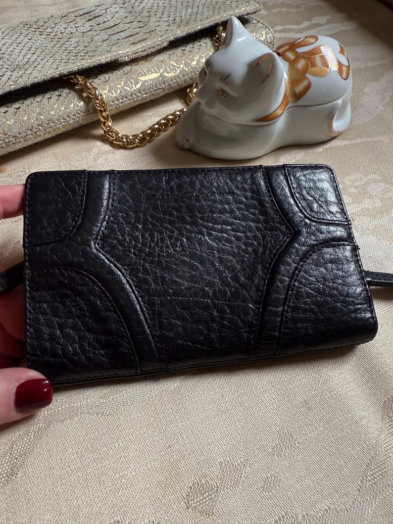 May include: Black leather wallet with a textured surface and a unique design. The wallet has a flap closure and a small strap.