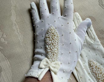 May include: A pair of off-white gloves with small pearl accents and a cluster of pearls on the back of the hand. A small bow is on the wrist. The gloves are made of a soft, textured fabric.