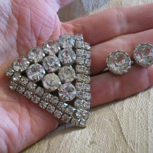 Pu&ograve; includere: Un fermaglio argentato con un grande gruppo triangolare di strass trasparenti. Il fermaglio &egrave; circondato da un bordo di strass pi&ugrave; piccoli. Un paio di orecchini abbinati con strass trasparenti.