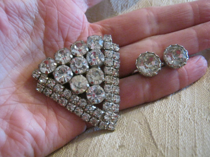 Pu&ograve; includere: Un fermaglio argentato con un grande gruppo triangolare di strass trasparenti. Il fermaglio &egrave; circondato da un bordo di strass pi&ugrave; piccoli. Un paio di orecchini abbinati con strass trasparenti.