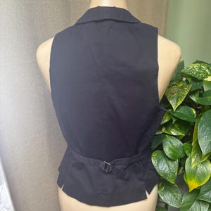 May include: Black vest with a notched collar and a belt with a silver-colored buckle. The vest is displayed on a mannequin. The back of the vest is visible, showing the tailored design and the adjustable belt.