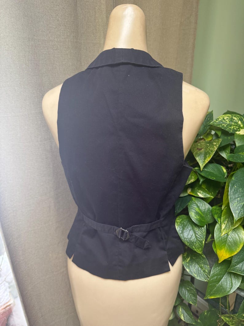 May include: Black vest with a notched collar and a belt with a silver-colored buckle. The vest is displayed on a mannequin. The back of the vest is visible, showing the tailored design and the adjustable belt.