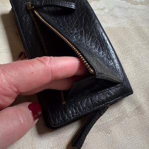 May include: A black leather wallet with a zipper closure and a small strap. The wallet is open and the inside is visible.