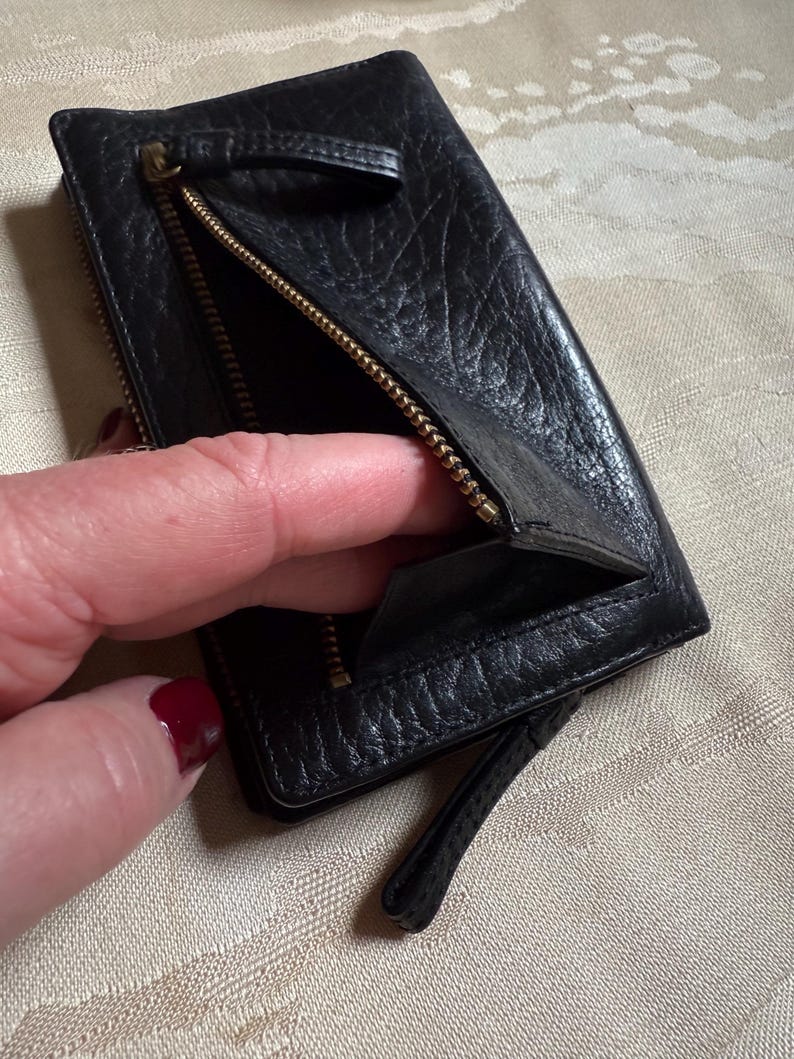 May include: A black leather wallet with a zipper closure and a small strap. The wallet is open and the inside is visible.