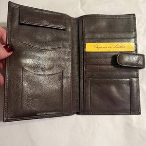 May include: Open brown leather wallet with multiple compartments and card slots. A yellow card with the text "Elegance in Leather" is visible. The wallet has a snap closure.
