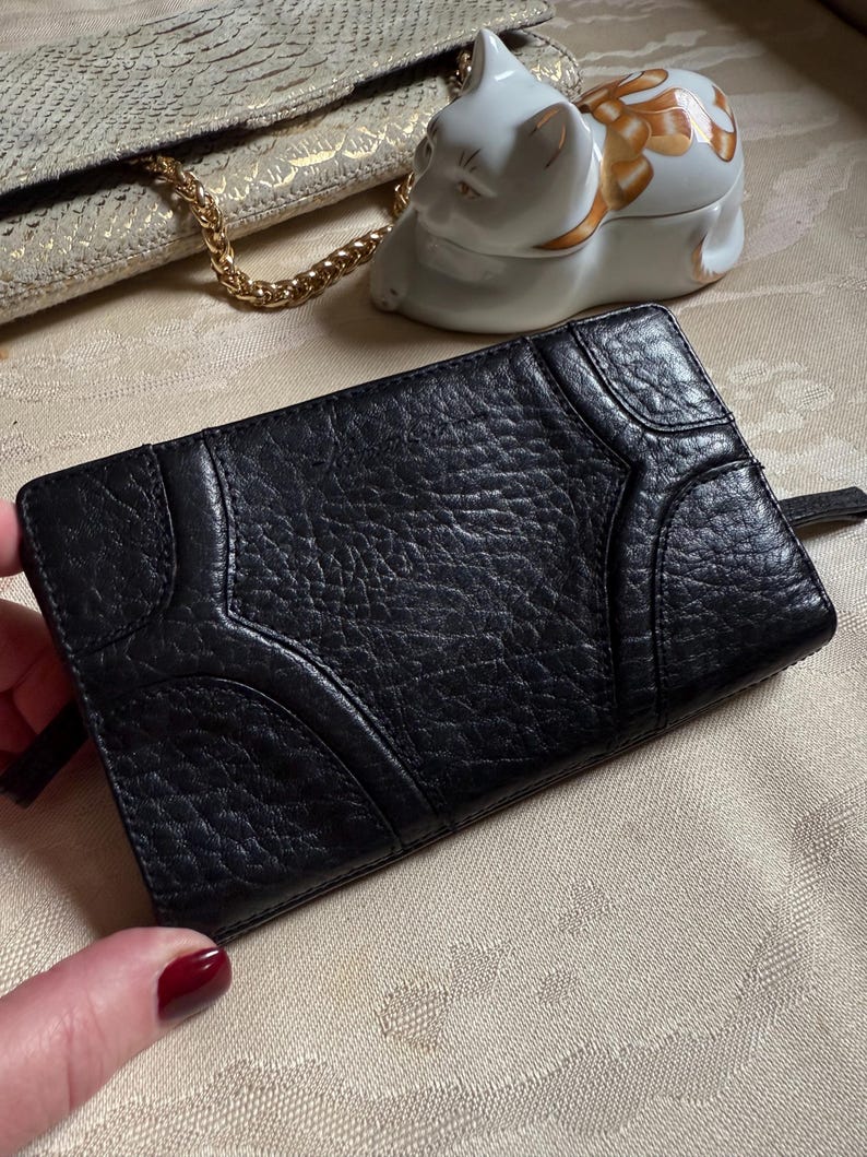 May include: Black leather wallet with a textured surface and a geometric design. The wallet has a zipper closure.