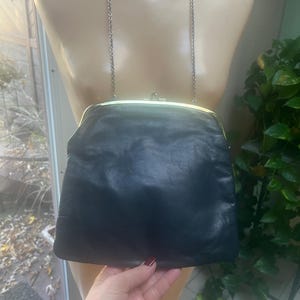 Pu&ograve; includere: Una pochette in pelle nera con una chiusura a scatto dorata e una tracolla a catena argentata.