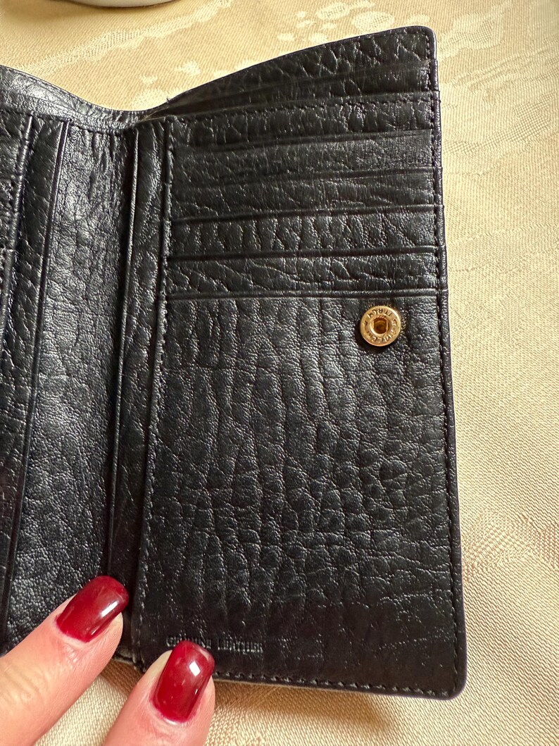 May include: Black leather wallet with multiple card slots and a snap closure. The wallet has a textured surface and a gold-colored button.