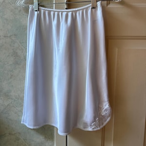 May include: A white satin half slip hanging on a clear plastic hanger. The slip has a smooth, shiny texture and a delicate lace trim along the hemline. The garment is designed to be worn under a skirt or dress.