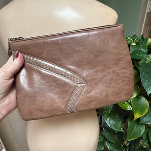 May include: A brown leather clutch with a snakeskin accent. The rectangular bag has a zipper closure and a wrist strap. A pearl necklace is draped over a mannequin.