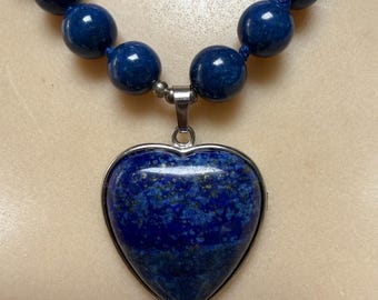 May include: A necklace featuring a series of round, deep blue lapis lazuli beads strung together, leading to a central heart-shaped lapis lazuli pendant encased in a polished silver-toned metal frame with a matching silver-toned metal bail.