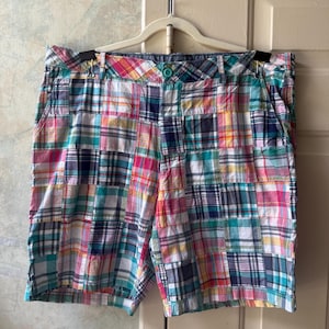 May include: These shorts feature a patchwork design composed of various plaid fabric squares in shades of pink, teal, yellow, navy blue, and white. The garment includes a waistband with belt loops, a single teal button closure, and side pockets.