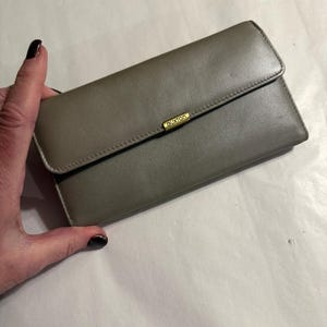 May include: A taupe-colored Buxton wallet with a rectangular shape and a fold-over flap. The wallet features a gold-toned metal accent with the brand name "Buxton" and is held by a hand with dark nail polish.