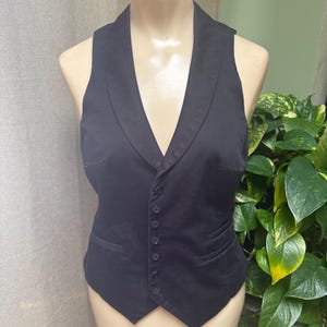 May include: A black vest with a shawl collar and a button-down front. The vest has a tailored fit and two front pockets. The garment is displayed on a mannequin. The background includes green plants and a neutral-colored curtain.