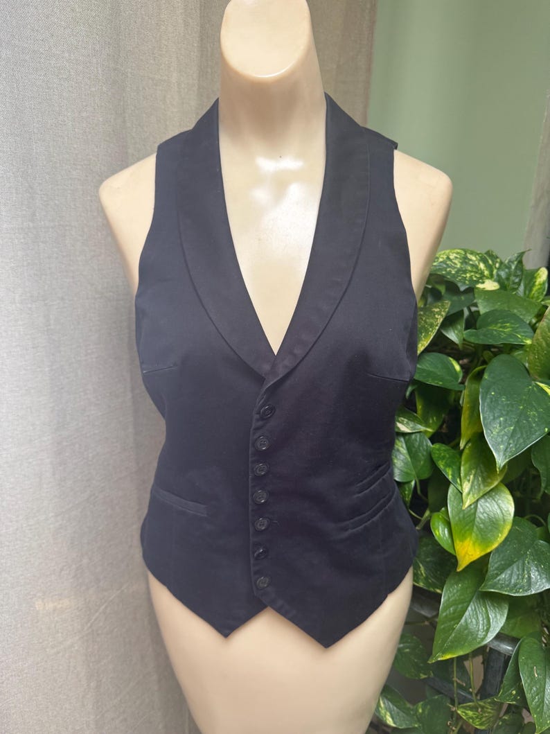 May include: A black vest with a shawl collar and a button-down front. The vest has a tailored fit and two front pockets. The garment is displayed on a mannequin. The background includes green plants and a neutral-colored curtain.