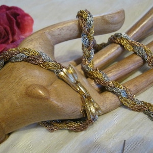 May include: A multi-tone necklace and bracelet set. The jewelry features a braided design with gold, silver, and copper-colored metal strands. The bracelet is fastened with a gold-toned clasp. A wooden hand prop holds the jewelry, with a red rose in the background.