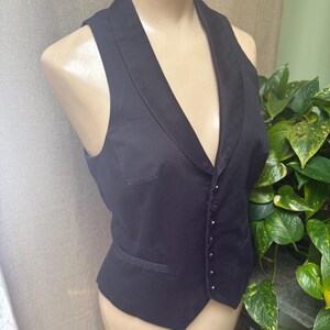 May include: A black vest with a shawl collar and a button-up front. The vest has a tailored fit and a small pocket on the left side. The garment is displayed on a mannequin.