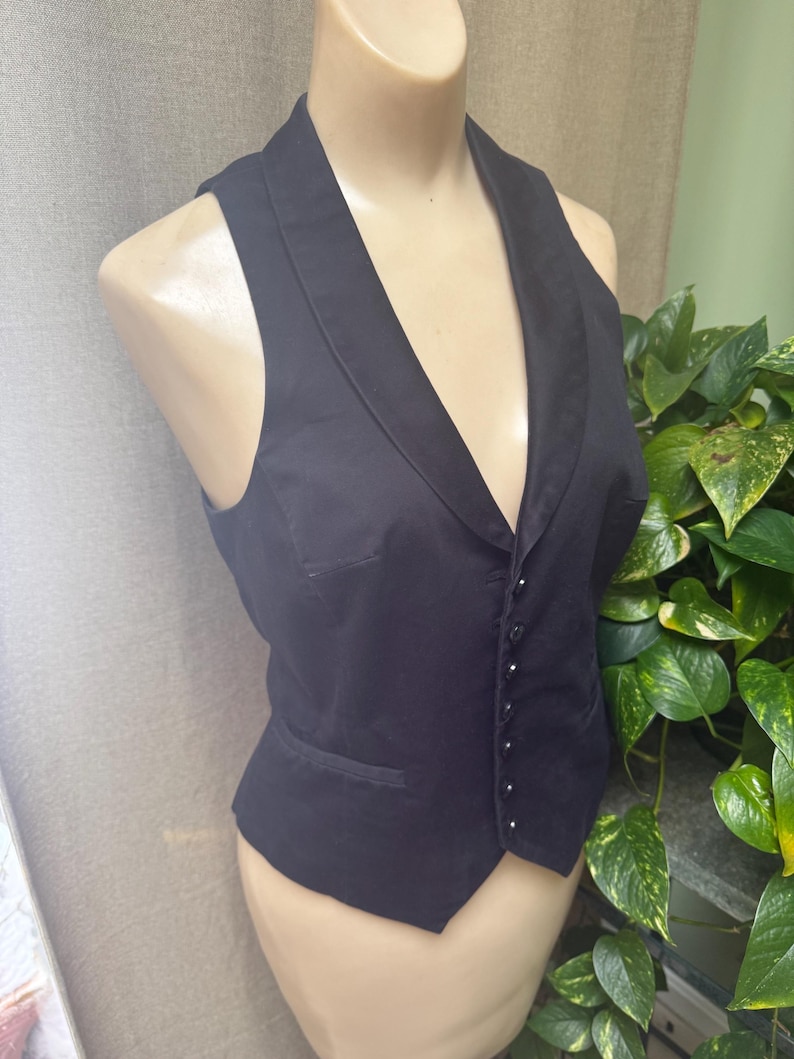 May include: A black vest with a shawl collar and a button-up front. The vest has a tailored fit and a small pocket on the left side. The garment is displayed on a mannequin.