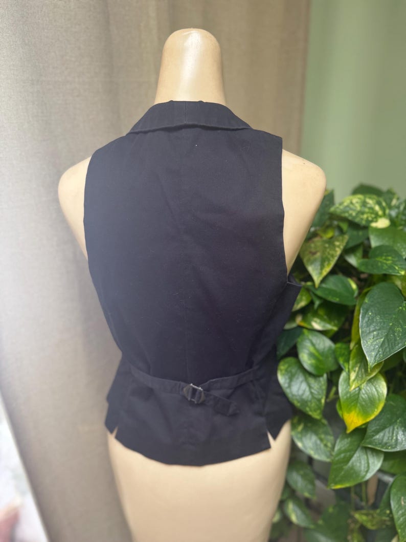 May include: Black vest with a collar and adjustable belt detail at the back. The vest is sleeveless and appears to be made of a woven fabric. The vest is displayed on a mannequin.