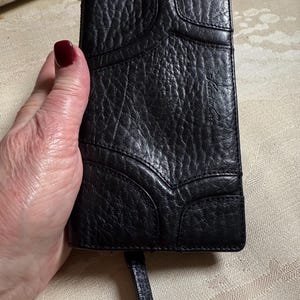 May include: Black leather wallet with a textured surface and a small strap. The wallet has a unique design with stitched panels.
