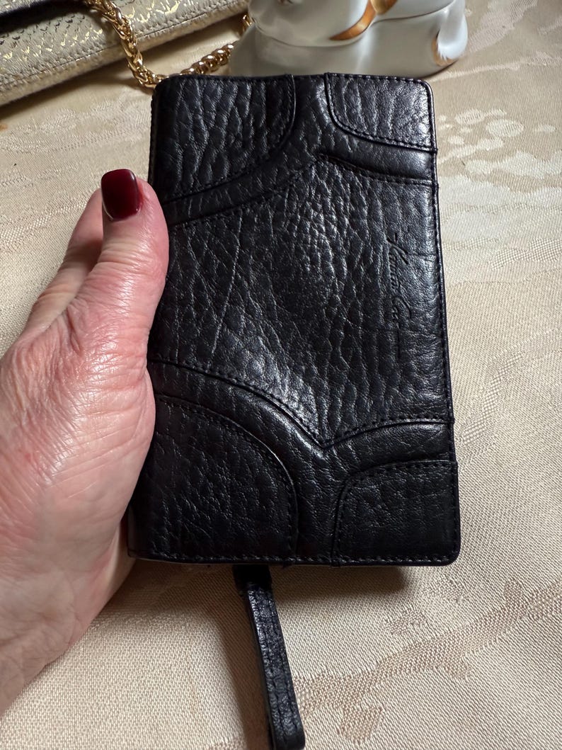 May include: Black leather wallet with a textured surface and a small strap. The wallet has a unique design with stitched panels.
