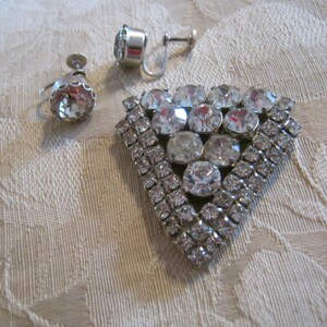 Pu&ograve; includere: Un set di gioielli vintage in strass con una spilla triangolare e un paio di orecchini a clip. La spilla &egrave; in metallo argentato e decorata con strass trasparenti di diverse dimensioni. Gli orecchini sono anch'essi argentati e presentano un unico grande strass trasparente.
