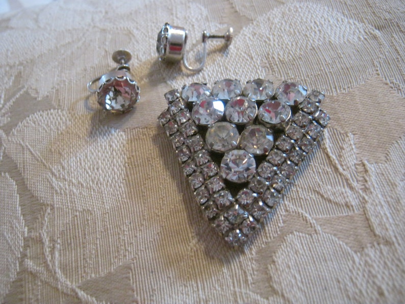 Pu&ograve; includere: Un set di gioielli vintage in strass con una spilla triangolare e un paio di orecchini a clip. La spilla &egrave; in metallo argentato e decorata con strass trasparenti di diverse dimensioni. Gli orecchini sono anch'essi argentati e presentano un unico grande strass trasparente.